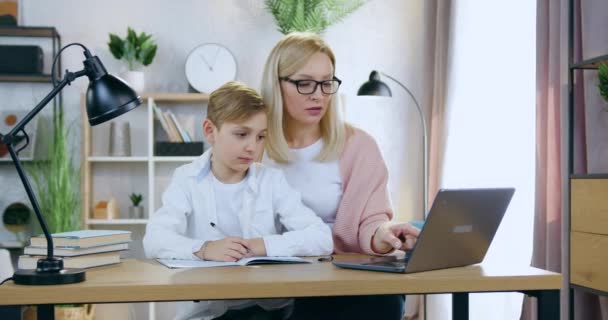 Portrét atraktivní zkušené starostlivé dospělé blond matky v brýlích, které sedí doma u stolu se svým krásným synem a pomáhá mu s on-line test, domácí vzdělávání koncepce — Stock video