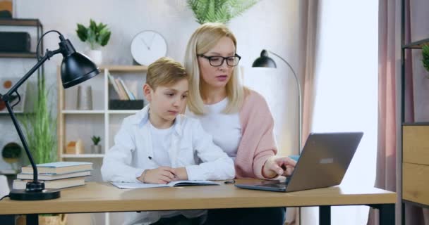 Retrato de atractiva madre rubia adulta hábil cuidadora en gafas que se sienta en el escritorio de casa con su hijo guapo y le ayuda con la prueba en línea, concepto de educación en el hogar — Vídeos de Stock