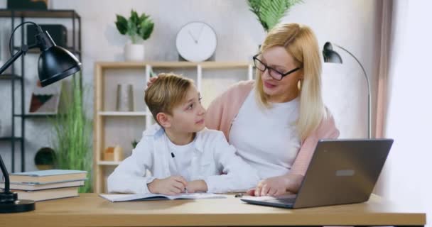 Muito feliz sorrindo 35-idade mãe de cabelos claros em óculos acariciando na cabeça seu atraente filho alegre ao ajudá-lo a fazer a tarefa de casa da escola na mesa no quarto acolhedor — Vídeo de Stock