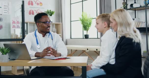 Bom aspecto positivo menino de 12 anos conversando com o médico sorridente de pele preta durante sua visita à clínica com adorável mãe loira satisfeita, pediatria e conceito de saúde — Vídeo de Stock