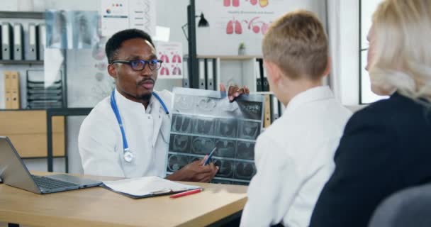 Yakışıklı, siyah tenli erkek doktor, modern klinikteki randevuları sırasında ergen hasta ve çekici konsantre annesine röntgen sonuçlarını açıklıyor. — Stok video