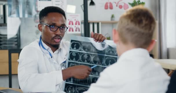 Bonito confiante hábil médico de pele preta em branco gawn explicando diagnisis de seu menino-paciente adolescente com mãe carinhosa usando meninos raio-x varredura durante sua visita ao consultório médico — Vídeo de Stock