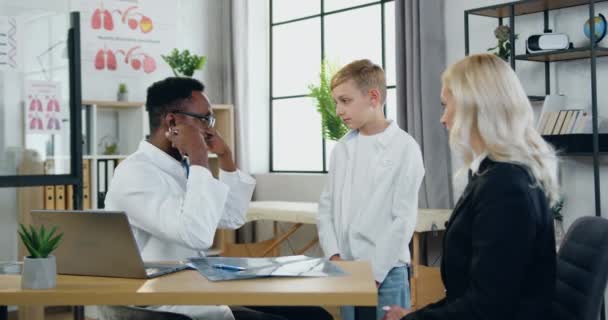 Attractive professional skilled african american doctor checking boys heartbeat during his scheduled visit to medical center with pretty caring loving mother — Stock Video