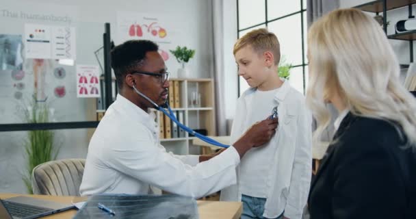 Healthcare and pediatry concept where likable confident skilled black-skinned male doctor checking boys lungs during his scheduled visit to clinic with beautiful mother — Stock Video