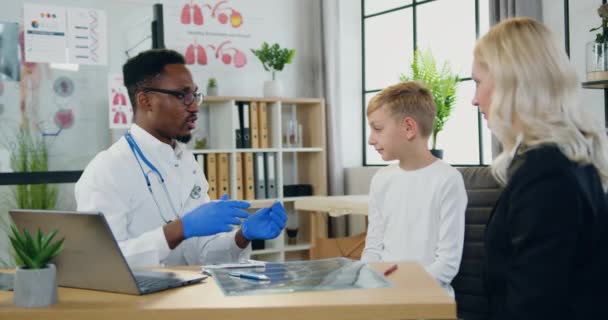 Adorável mulher carinhosa assistindo como profissional amigável afro-americano masculino médico examinando seus filhos adolescentes garganta com espátula de madeira durante sua visita à clínica — Vídeo de Stock