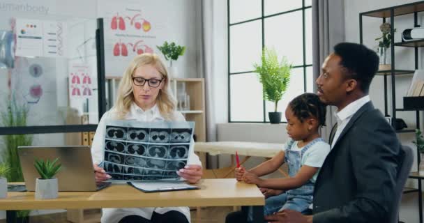 Hermosa profesional positiva rubia mujer médico explicando las niñas imagen de rayos X del cerebro a guapo padre de piel negra serio cuidado durante la visita programada con su hija al centro médico — Vídeo de stock