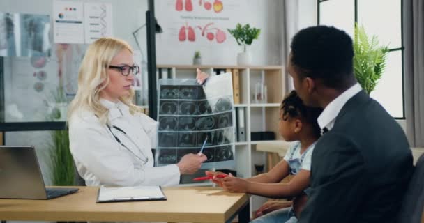 Çekici, dost canlısı, deneyimli sarışın kadın-doktor, kliniğe gelip röntgen sonuçlarını öğrenirken, yakışıklı, şefkatli, siyah tenli ve küçük kızı olan bir adamla konuşuyor. — Stok video