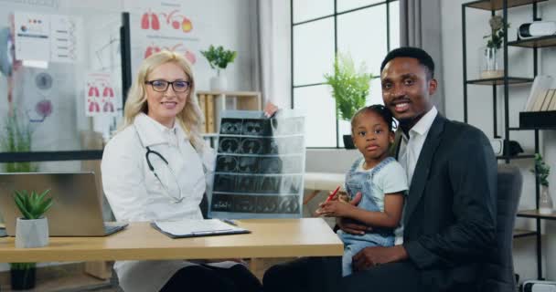 Adorable uśmiechnięty doświadczony blond kobieta pediatra trzymając rentgenowskie skanowanie i gesting kciuki do góry wraz z małym cute girl-pacjent i jej przystojny wesoły zadowolony czarny skóry ojciec w — Wideo stockowe