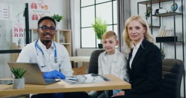 Sağlık sigortası konsepti. Yakışıklı, gülümseyen siyah tenli doktor, oldukça olumlu bir kadın ve ergenlik çağdaş klinik ofisinde kameraya işaret eden oğlu.