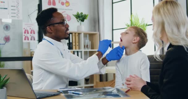 Knappe positieve hoogopgeleide zwarte huid lokale arts is tevreden algemene inspectie van jongens keel tijdens geplande bezoek aan zijn eigen medische kantoor samen met glimlachende gelukkige moeder — Stockvideo