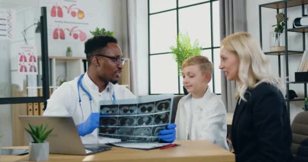 Goed uitziende glimlachende vriendelijke behendige zwarte huid arts is tevreden van x-ray beeld van zijn jongen-patiënt en het geven van high five elkaar tijdens zijn bezoek in kliniek met schattige gelukkige moeder — Stockvideo