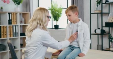 Çekici, kendine güvenen, deneyimli sarışın kadın-doktor kalp atışlarını ve akciğerlerini kontrol eden modern tıp ofisinde rahat genç bir çocuğun, sağlık konsepti.