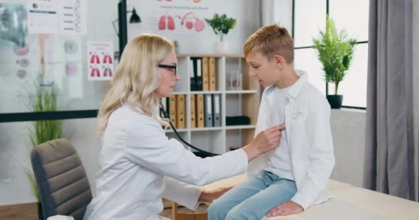 Imádnivaló mosolygós elégedett képzett női gyermekorvos vizsgálja szívverés és a tüdő pozitív 12 éves fiú a tervezett látogatás a klinikán, és beszélni vele ellenőrzés után — Stock videók