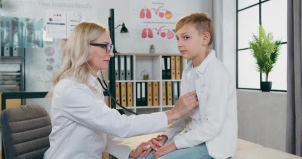 Великолепная улыбающаяся высококвалифицированная блондинка-врач проверяет сердце и лёгкие мальчиков в современном медицинском кабинете, и они смотрят в камеру с поднятыми пальцами — стоковое видео