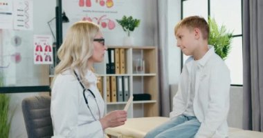 Modern klinik ve pediatri konseptine yapacağı ziyarette erkek çocukların boğazlarını ve ağızlarını kontrol eden güzel, dost canlısı sarışın çocuk doktoru.