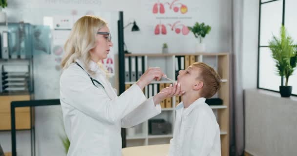 Piękny profesjonalny pozytywny pozytywny blondynka lekarz badając chłopców gardła z łopatką podczas genaral inspekcji w nowoczesnym gabinecie medycznym — Wideo stockowe