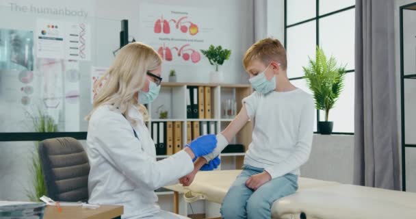 Concepto de atención médica donde el cuidado atractivo bondadoso médico rubio mujer imponiendo vendaje apretado en la mano de los niños en el gabinete médico moderno — Vídeo de stock