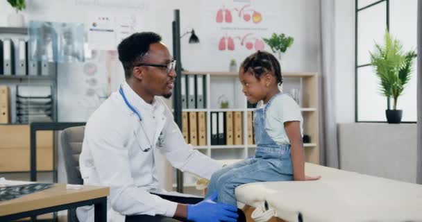 Boa aparência sorridente carinhoso afro-americano médico falando com a menina alegre no escritório médico moderno e dando-lhe alta cinco, conceito de medicina — Vídeo de Stock