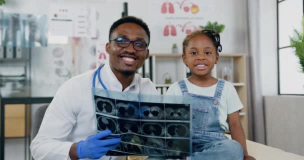 Vue de face de beau souriant soignant hautement qualifié à la peau noire médecin masculin qui regarde la caméra avec une petite fille noire joyeuse et tenant dans ses mains les résultats des rayons X — Video