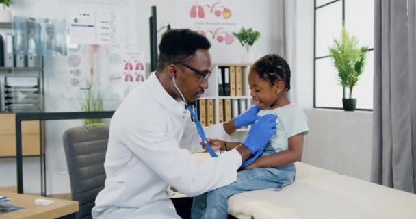 Probable cuidador sonriente pediatra masculino de piel negra que examina los latidos cardíacos de las niñas con estetoscopio y después de terminar la inspección que le da cinco altos — Vídeos de Stock