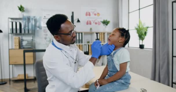 Красивый уверенный в себе профессиональный чернокожий мужчина-врач осматривает горло маленьких девочек лопаткой в своем медицинском кабинете в современном центре лечения — стоковое видео