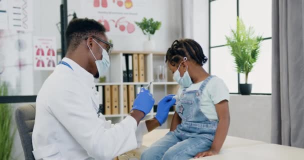 Atractivo profesional habilidoso médico varón de piel negra en máscara facial haciendo inyección en el brazo de las niñas pequeñas durante su visita a la clínica, concepto de salud — Vídeos de Stock