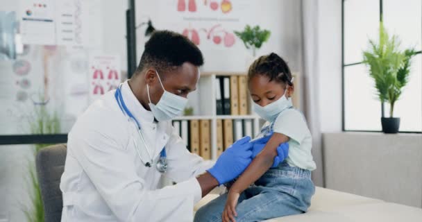 Beautiful 6-aged black-skinned girl in facial mask visiting her local experienced african american male doctor and watching how he doing injection in her arm — Stock Video