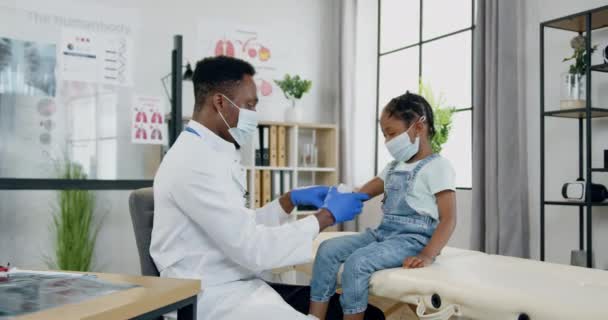 Bella calma dalla pelle nera piccola ragazza in maschera facciale seduta sul tavolo di revisione in studio medico mentre bello premuroso medico maschio dalla pelle nera professionale in maschera protettiva imponendo benda su di lei — Video Stock