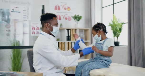 Nice calma pequena menina negra em máscara facial sentado na mesa de revisão no consultório médico e assistindo como atraente experiente cuidar de pele preta médico masculino em máscara protetora impondo apertado — Vídeo de Stock