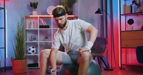 Ελκυστικός δραστήριος κουρασμένος νεαρός γενειοφόρος άνδρας στο αθλητικό ντύσιμο κάθεται σε fitball και κάνει ασκήσεις με αλτήρες για να κρατήσει το σώμα του υγιές και ισχυρό, μπροστινή άποψη — Αρχείο Βίντεο