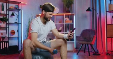 Sağlıklı yaşam tarzı konsepti. Alıştırma kıyafetleri içinde çekici, sportif, genç bir sakallının evde telefonuyla komik videolar izlemesi.