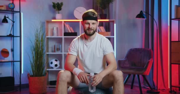 Portrait of good-looking smiling sporty young bearded guy in training clothes whic showing a bottle with water when doing home workout in the evening — Stock Video