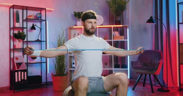 Goed uitziende actieve sportieve jonge bebaarde man in sportkleding zittend op fitbal thuis in de avond en het doen van oefeningen met stretching weerstand band — Stockvideo