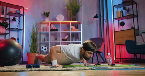 Концепция выносливости дома тренировки, где красивый сильный активный современный бородатый парень в спортивной одежде делает отжимания упражнения на полу в вечернее время дома — стоковое видео