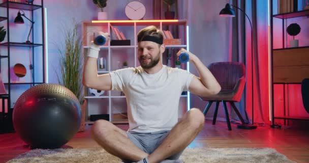 Síla doma cvičení koncept, kde sympatický úsměv sportovní vousatý muž ve sportovním oblečení sedí v lotosu pózovat na podlaze a cvičení s činky — Stock video