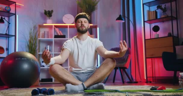 Guapo chico barbudo joven relajado en ropa deportiva sentado en la pose de loto en el suelo en casa y meditando con los ojos cerrados para disfrutar del silencio y la armonía a su alrededor — Vídeos de Stock