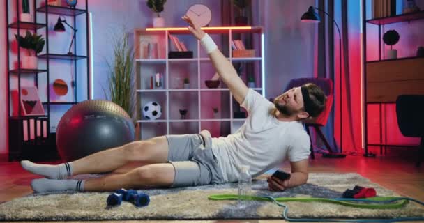 Dobře vypadající pozitivní trochu unavený vousatý sportovec ležící na boku na podlaze a dělá cvičení s rovnající se nohou během domácího cvičení — Stock video