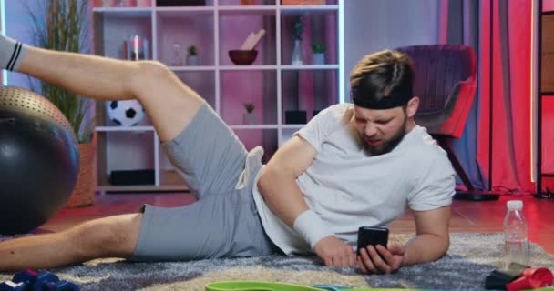 Přední pohled na dobře vypadající sportovní silný mladý vousatý chlap ve sportovním oblečení, které dělá cvičení pro nohy ležící na podlaze doma ve večerních hodinách současně sledování videa na telefonu — Stock video