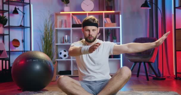 Portret dobrze wyglądającego spokojnego, wyszkolonego brodatego faceta w odzieży sportowej, który wieczorem siedzi w lotosowej pozycji na podłodze w domu i wykonuje relaksujące ćwiczenia jogi — Wideo stockowe