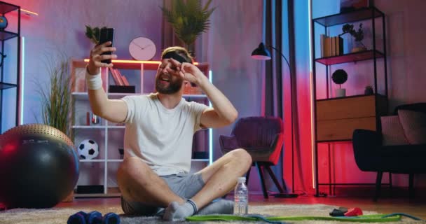 Aantrekkelijk lachend ontspannen jonge bebaarde sportman in trainingskleding zittend met gekruiste benen op de vloer thuis en grappige selfies makend op zijn smartphone — Stockvideo