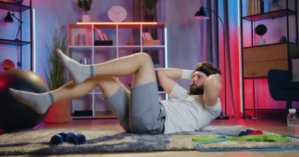ボディケアのコンセプトここで魅力的なアクティブスポーティひげそり男でスポーツウェアでやってcrissrossクランチ演習で床に自宅で夜 — ストック動画