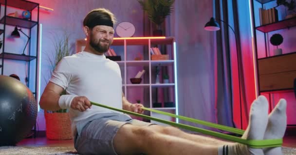 Концепция домашней тренировки, где симпатичный спортивный босоногий мужчина в спортивной одежде делает упражнения с резиновым ремнем на полу дома вечером — стоковое видео