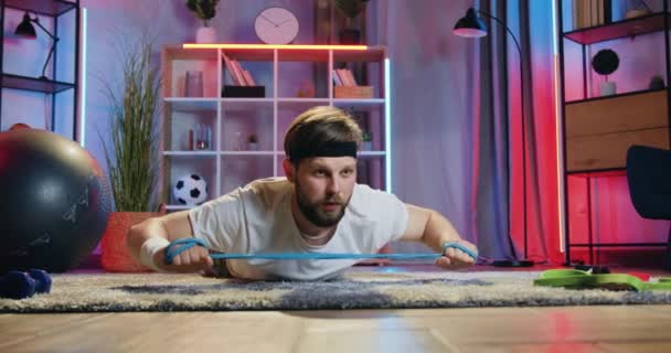 Концепция здорового образа жизни, где привлекательный концентрированный активный мужчина в спортивной одежде лежит на полу дома и делает упражнения на растяжку с эластичной лентой — стоковое видео