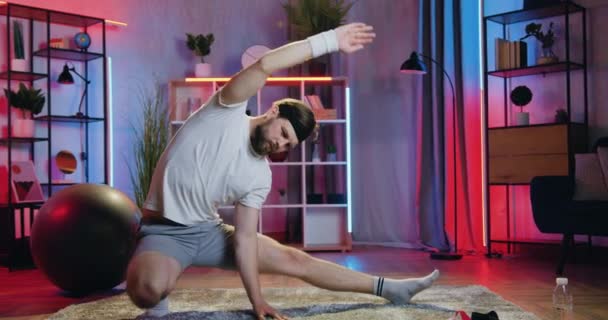 Prawdopodobnie aktywny sportowy brodaty facet siedzi squat na podłodze w przytulnym pokoju w godzinach wieczornych i robi ćwiczenia rozciągające ramiona, koncepcja treningu w domu — Wideo stockowe