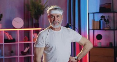 Sevimli, güçlü, sakallı, spor kıyafetli bir adam kameranın önünde duruyor ve ellerini göstererek iç mimarinin arka planında kol kaslarını gösteriyor.