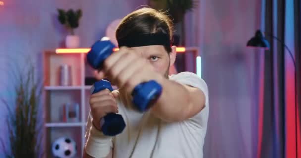 Portrait of likable active young bearded sportsman in white t-shirt and black headband which work outing his boxing punches with dumbbells in his hands at home — 图库视频影像