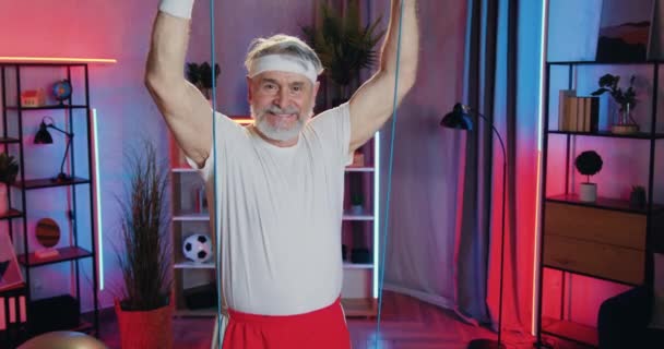Sympatický úsměv aktivní sportovní starý vousatý muž ve sportovním oblečení dělá sílu cvičení pomocí odporu gumičky během domácího večerního cvičení — Stock video