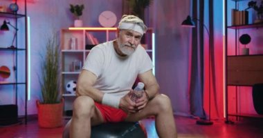 Spor giyim sektöründe sportif ve olgun sakallı bir adamın portresi. Ev antrenmanından sonra spor salonunda otururken kameraya iyi bir sembol gösteriyor.