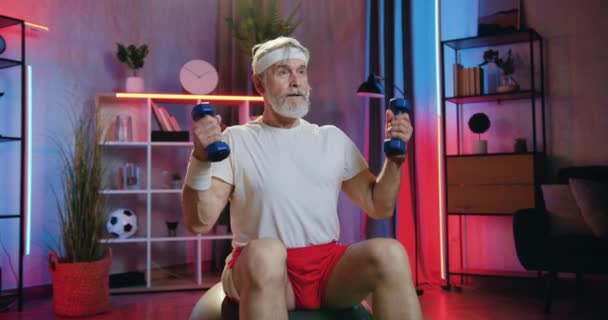 Вероятный уверенный в себе активный старший бородатый мужчина в спортивной одежде, сидя на фитболе и тренируясь с гантелями во время домашней вечерней тренировки, закрыть — стоковое видео
