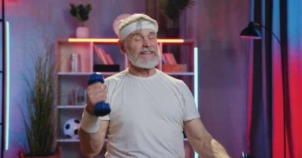 Красивый активный спортивный бородатый старик в спортивной одежде интенсивно занимается спортивными упражнениями с гантелями во время домашней вечерней тренировки, вид спереди — стоковое видео